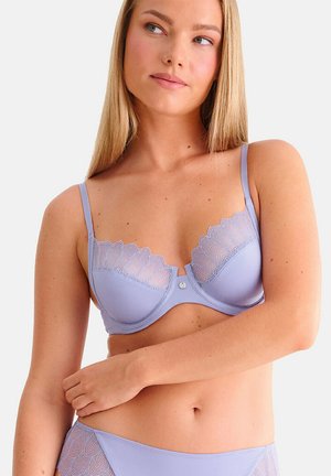 Femme portant un soutien-gorge violet clair orné de dentelle et une culotte assortie, posant les bras croisés, regardant sur le côté sur un fond uni.
