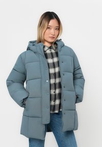 Pūtuvs jaku mīkstajā zilo krāsā ar kapuci, ar kvadrātveida šuvēm un spiedpogu aizdari. Nēsāta virs pārbaudešādas krekla un džinsu biksēm.