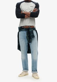 Graues und marineblaues Langarmhemd, blaue Jeans und braune Sneakers. Dunkelgrünes Flanellhemd um die Taille gebunden. Brauner Gürtel und Uhr.