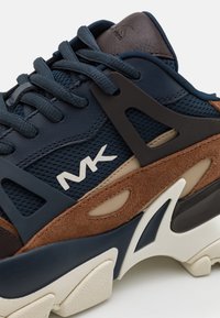 Zapatilla de deporte azul marino, marrón y beige con paneles de malla, ante y cuero, suela gruesa y cordones azules marinos, con el logo "MK" en el lateral.