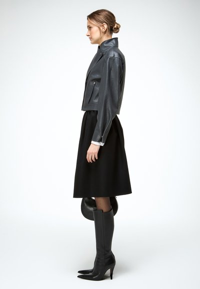 Vrouw staand in profiel, gekleed in een cropped zwarte leren jas, zwarte midi rok, zwarte hoge knielaarzen met hakken, en houdt een ronde zwarte tas vast.