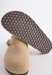 Sabots en daim beige avec un bout arrondi, dotés d'une bride à boucle unique et d'une semelle en caoutchouc à motif alvéolé pour une meilleure traction.