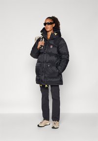 Black puffer jacket with a hood, featuring yellow drawstrings and front pockets. Worn with dark cargo pants and beige hiking shoes.