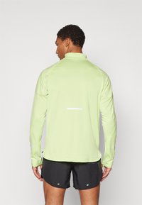 Hellgrüne langärmlige Sportoberteil mit Stehkragen und reflektierenden Details. Getragen mit schwarzen Shorts, die ein figurbetontes Design und strukturierten Stoff zeigen.