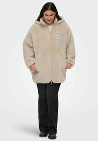 Bežs flīsa kapucei sporta jaka ar rāvējslēdzēju priekšpusē, plaša piegriezuma, sānu kabatām un krūšu kabatu. Mīksta tekstūra un bieza materiāla.