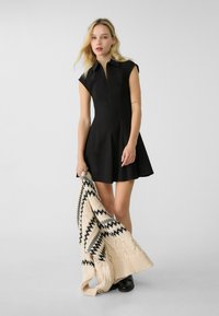 Sort kjole til knæet med tætsiddende overdel og cap sleeves, parret med et cremefarvet strikket sjal med sorte geometriske mønstre.