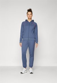 Marineblå treningsdrakt med rosa detaljer. Inkluderer en glidelåshettejakke og tapered bukser, laget av et mykt, elastisk stoff.