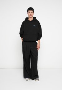 Hanorac supradimensionat negru cu buzunar frontal și logo mic albastru, asortat cu pantaloni negri largi cu model în carouri și încălțăminte casual.