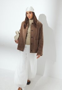 Brun ruskindsjakke med en afslappet pasform, der har knaplukning og to frontlommer. Bæres over en beige bluse og hvide brede bukser.