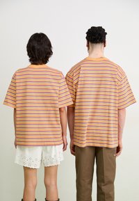 Svītraini pārmērīgi T-krekli oranžā, dzeltenā un violetā krāsā ar īsām piedurknēm, apvienoti ar baltām viļņotām bermudām un brūnām biksēm.