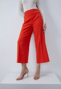 Pantalon ample rouge vif en tissu lisse, à taille haute et lignes épurées, associé à des talons hauts nus à lanières.
