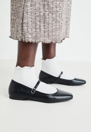 Mustad musta baleriinna, millel on terav varvas ja tasane konts, ning reguleeritav rihm jala üle. Kanda koos valgete tekstuursete sokkidega.