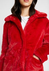 Rød faux fur frakke med en fyldig tekstur, krave og drawstring i taljen. Har sidelommer og knaplukning for praktisk brug.