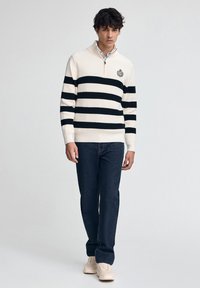 Strick-Polo-Sweater in Creme mit marineblauen horizontalen Streifen, Reißverschluss-Kragen und besticktem Logo. Kombiniert mit dunkelblauer Jeans und Sneakers.