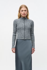 Pulover gri cu fermoar și model zimțat, guler înalt și mâneci lungi, asortat cu o fustă gri netedă, pe un fundal alb simplu.