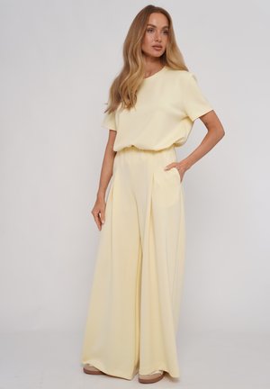 Kobieta o długich blond włosach, ubrana w bladożółty krótki top z krótkim rękawem i szerokie spodnie z wysokim stanem, stoi z jedną ręką w kieszeni.