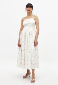 Vestido maxi blanco con flores, con tirantes finos, cintura fruncida y falda fluida. Presenta patrones de flores coloridos en rosa, amarillo y verde.