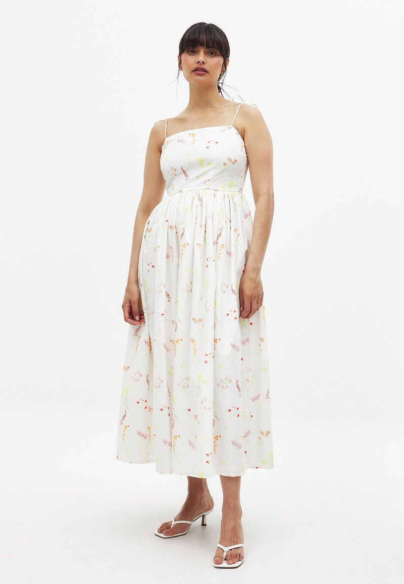 Vestido maxi blanco con flores, con tirantes finos, cintura fruncida y falda fluida. Presenta patrones de flores coloridos en rosa, amarillo y verde.