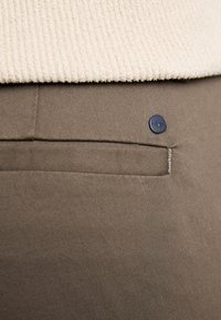 Gros plan sur la poche d'un pantalon marron avec une étiquette de marque noire circulaire et l'ourlet d'un pull beige texturé au-dessus.