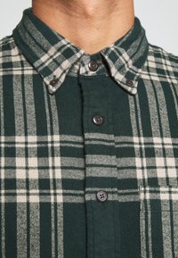 Groen geruit flanellen overhemd met een button-down kraag, voorzien van lichtbeige en donkergroene strepen, en bruine knopen. Zware textuur.
