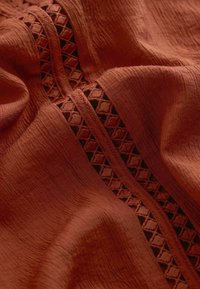 Rust-colored fabric with a textured, crinkled surface and a decorative geometric mesh stitched in a diagonal pattern.