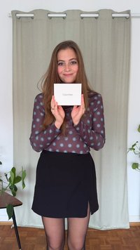 Calvin Klein hvit eske holdt av en person i brun langermet skjorte med blå prikker og svart skjørt, mot en lys bakgrunn.