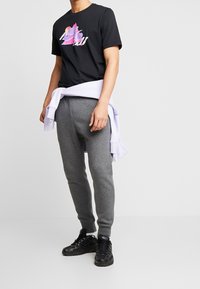 Svart t-shirt med kort ärm och grafisk logotyp i rosa och lila, tillsammans med ljusgråa träningsbyxor och svarta sneakers. Vit sweatshirt knuten i midjan.