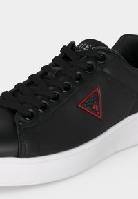 Μαύρο δερμάτινο αθλητικό παπούτσι με μαύρα κορδόνια, λευκή σόλα και κόκκινο τριγωνικό λογότυπο Guess στην πλαϊνή πλευρά.