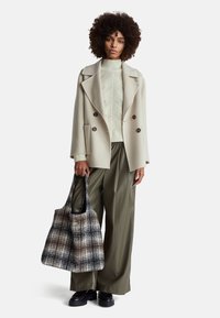 Kremowy płaszcz dwurzędowy noszony na teksturowanym jasnym swetrze, w połączeniu z oliwkowymi spodniami o szerokich nogawkach i w kratkę torbą w stonowanych odcieniach.