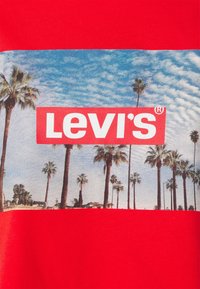 T-shirt rouge avec un graphisme de palmiers sur fond de ciel bleu, arborant un grand logo blanc "LEVI'S" dans un encadré rouge. Texture en tissu doux.