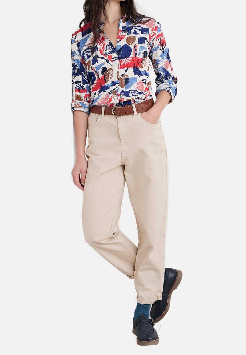 Femme portant une chemise à motifs colorés boutonnée, pantalon beige taille haute avec ceinture marron, chaussettes bleues et mocassins noirs, debout avec une main dans la poche.