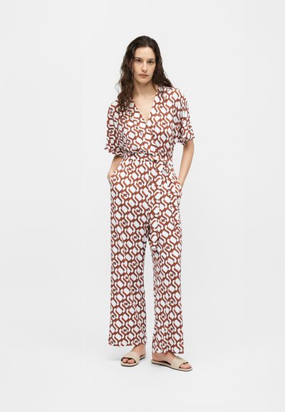 Γυναίκα με μακριά κυματιστά μαλλιά φοράει καφέ και άσπρη ολόσωμη φόρμα με κοντά μανίκια, τσέπες, φαρδιά πόδια και μπεζ σανδάλια slide.