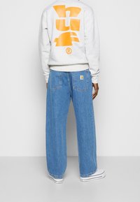 Sudadera gris claro con gran logo naranja "huf" en la parte posterior, combinada con unos vaqueros de denim azul de corte holgado que cuentan con un parche de logo en el bolsillo trasero.