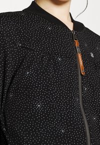 Svart bomberjacka med ett fläckigt mönster av små grå prickar, med en ribbad krage och en metalldragkedja med en läderdragare.