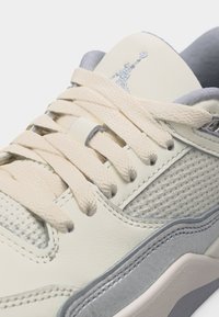 Κοντινό πλάνο μπεζ και γκρίζου αθλητικού παπουτσιού με υφασμάτινα κορδόνια, πλευρική διχτυωτή επιφάνεια και κεντημένο λογότυπο Jumpman στη γλώσσα.