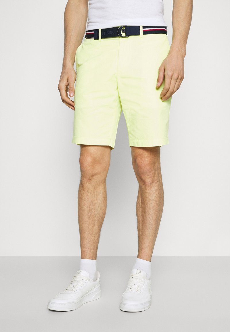 Homme portant un short jaune clair, des baskets blanches, des chaussettes blanches, une ceinture noire avec des rayures rouges et blanches, et un t-shirt blanc.