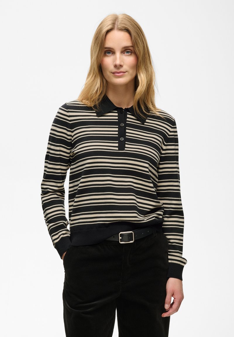 Femme aux cheveux blonds portant un polo à manches longues rayé noir et beige et un pantalon noir, les mains dans les poches, sur un fond uni.