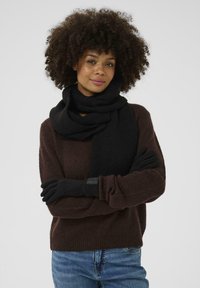 Schwarzer gestrickter Schal um den Hals drapiert, kombiniert mit schwarzen Handschuhen. Das Modell trägt einen braunen Pullover. Die Textur wirkt weich und warm.