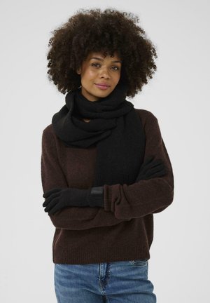 Schwarzer gestrickter Schal um den Hals drapiert, kombiniert mit schwarzen Handschuhen. Das Modell trägt einen braunen Pullover. Die Textur wirkt weich und warm.