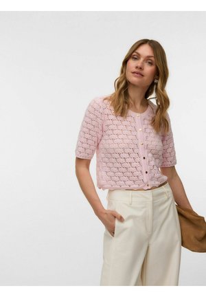 Femme portant un cardigan en tricot rose à manches courtes avec des boutons et un pantalon crème taille haute, tenant une pochette marron dans sa main gauche.