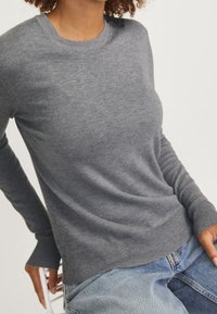 Grå stickad tröja med lång ärm och rundad halsringning, ribbade muddar, matchad med blå denimjeans, visad på en sittande figur.