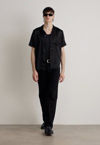 Maglietta nera a maniche corte con righe verticali, abbinata a una canottiera nera, pantaloni e scarpe. Gli accessori includono occhiali da sole scuri.