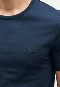 Marineblaue T-Shirt mit Rundhalsausschnitt und kurzen Ärmeln mit "BOSS" auf der linken Brust bestickt, getragen von einer Person.