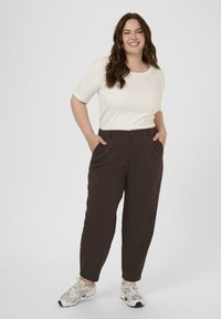 Brune bukser med lige ben-design, der har sidelommer og en frontknap. Sammen med en cremefarvet skjorte med korte ærmer og sneakers.