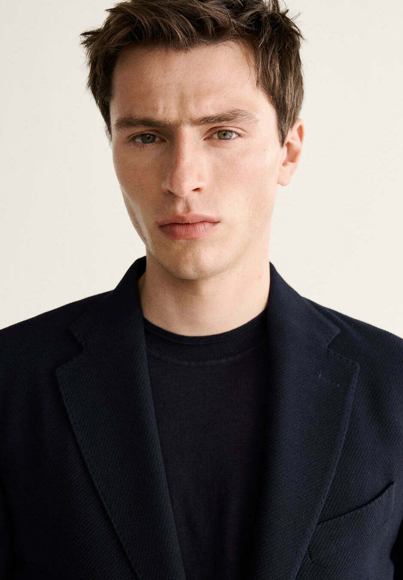 Joven con cabello castaño corto, vistiendo un blazer negro y una camisa negra, mirando directamente con una expresión neutral sobre un fondo liso.