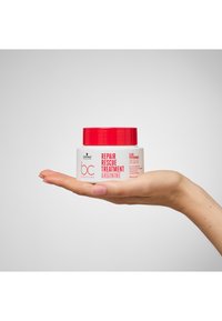 Contenitore rotondo bianco con coperchio rosso, etichettato "Trattamento di Riparazione Rescue Arginina". Tenuto in mano contro uno sfondo neutro.