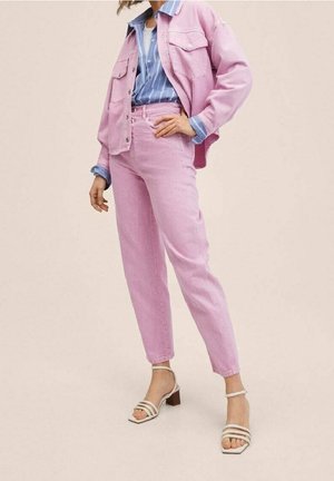 Kvinde iført lyserød denimjakke og bukser over blåstribet skjorte, poserer med hånden på hoften, stående i beige sandaler med blokhæl.