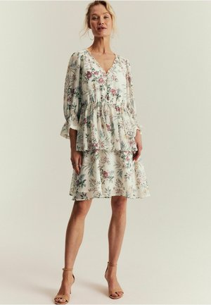 Frau trägt ein weißes, knielanges Kleid mit Blumenmuster, langen Ärmeln und Rüschendetails, steht vor einem schlichten hellen Hintergrund.