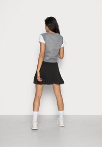 Vrouw staat met haar rug naar de camera, draagt witte sneakers, witte sokken, een zwarte geplooide rok, een wit shirt met korte mouwen en een grijze mouwloze vest.