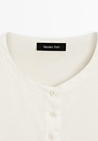 Krémová bavlněná košile s kulatým výstřihem a knoflíkovým zapínáním. Obsahuje černou nálepku s nápisem "Massimo Dutti." Hladká textura.